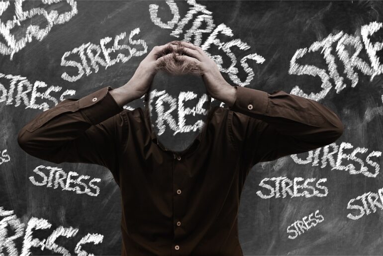 Mann schlägt Hände über seinem Kopf zusammen, dahiner befindet sich eine Tafel mit den Aufdrucken Stress